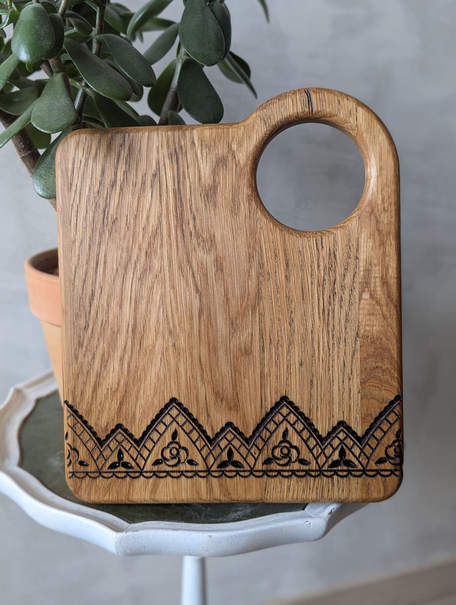 Handgefertigtes Eichen-Schneidebrett mit rundem Griff und laser-graviertem geometrisch-floralem Bortenornament am unteren Rand